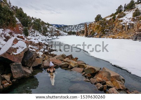 Cheerful young Caucasian woman in a black bikini lies in the therapeutic water of Radium hot springs and observes the beautiful snowy landscape. Happy hiker girl relaxes in a natural spa in Colorado. Royalty-Free Stock Photo #1539630260