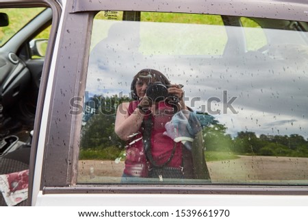 The girl with the camera is reflected in the glass of the car. Photographer takes pictures on a summer trip by car