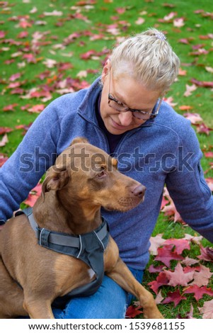 Doberman mix dog sitting in blond woman’s lap, outside on green lawn covered in red fall leaves
