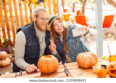 Young couple man and woman sitting at table outdoors at vacation home making jack-o'-lantern preparing for halloween taking selfie photo on smartphone posing to camera smiling cheerful