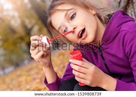 Little girl sitting under the tree outdoors in the autumn park blowing soap bubbles looking at bubble smiling surprised