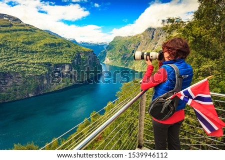 Tourism holidays picture and traveling. Woman tourist enjoying fjord landscape Geirangerfjord from Ornesvingen eagle road viewpoint, taking photo with camera, Norway.