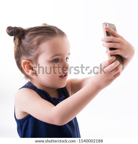 Portrait of cool cheerful little girl taking photo with smart phone. Cute kid shooting selfie on front camera isolated on white background. Child using a mobile phone.