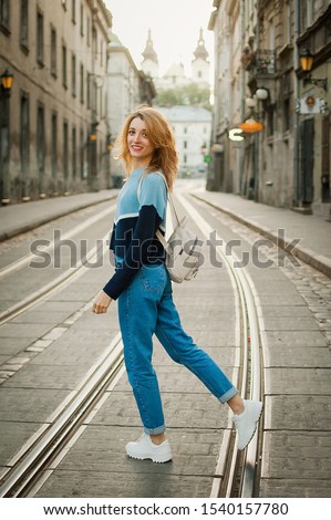 Full height portrait of young woman in blue casual sweater, jeans, white shoes, and small backpack early in the morning in ancient city in Europe on empty street with tramway background.
