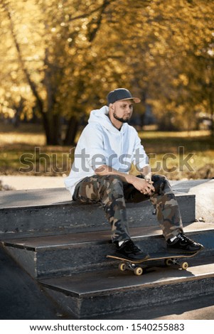 Man with beard in skatepark with skateboard sits on the stairs on warm autumn day