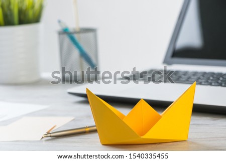 Businessman workspace with office wooden desk and yellow paper ship. Flat lay table with plant in pot and laptop computer. Close up orange origami boat. Creative and innovative solution for business.
