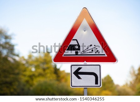 A triangular red white warning sign. The road is under construction and there are small stones and granules on the road. In the background, green trees out of focus with beautiful bokeh.