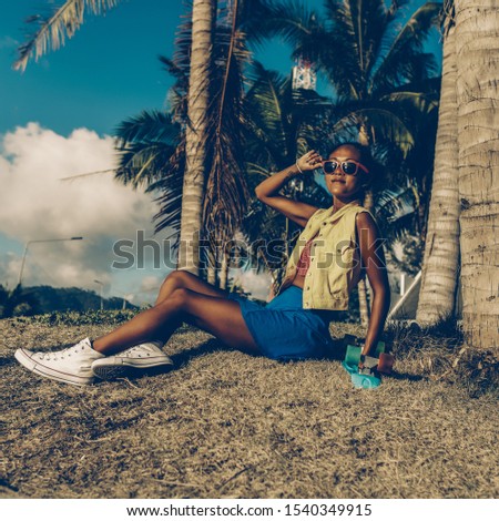 Outdoor lifestyle portrait of beautiful black lady in bright outfit and white sneakers. Hipster woman sit at dry grass under palm tree and touch her sunglasses. Sunny summer hot day. Swag, fashion.