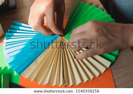 In the foreground female hands glue origami fans, sheets of colored paper, scissors on a wooden table. Several blanks for the fan. Template for design, advertising or text.