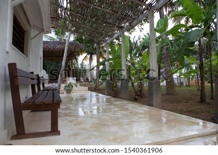 day shot of a terrace of a house with a wooden bench inside a tropical garden during a heavy rain storm with tall wet palm and banana trees and their large green leaves and water everywhere. Sri Lanka