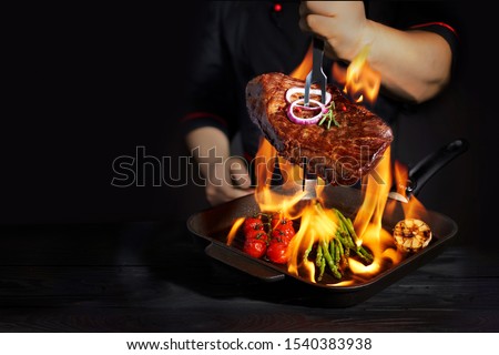 Chef at the fork to cook a steak and vegetables in pan with fire flame on  dark background. Restaurant and hotel service concept. Royalty-Free Stock Photo #1540383938