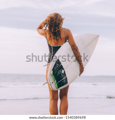 Surf girl with long hair go to surfing. Young surfer woman holding blank white short surfboard on a rock at sunset or sunrise. Bali island, Indonesia. Outdoor Active Lifestyle. It's time for surfing!