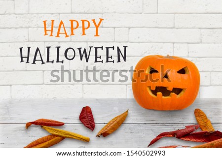Banner Happy Halloween with pumpkin jack lantern standing on white wood in front of white brick wall, text in black and orange, autumn leaves around
