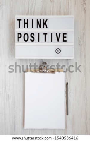 'Think positive' words on a modern board, clipboard with blank sheet of paper on a white wooden surface, top view. Overhead, from above, flat lay. Close-up.