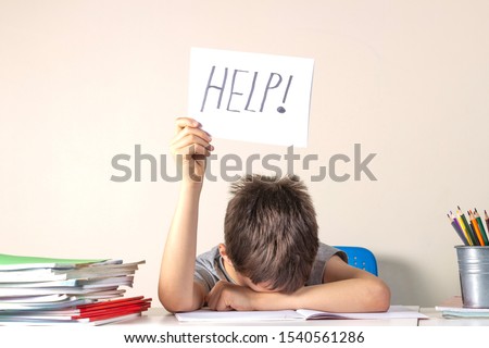 Sad tired frustrated boy sitting at the table with many books and holding paper with word Help. Learning difficulties, education concept. Royalty-Free Stock Photo #1540561286