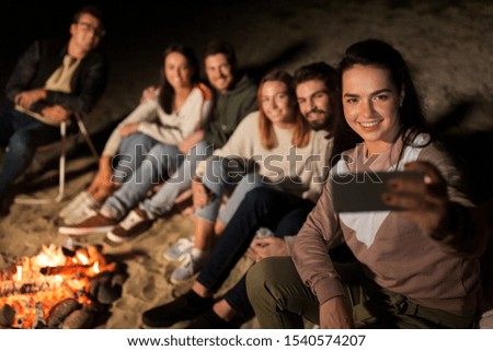 leisure and people concept - group of smiling friends sitting at camp fire taking selfie by smartphone on beach at night
