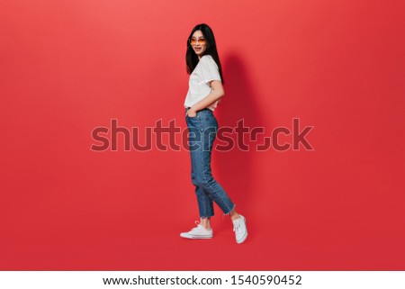 Lady in great mood is posing on red background. Portrait of Asian in jeans and T-shirt