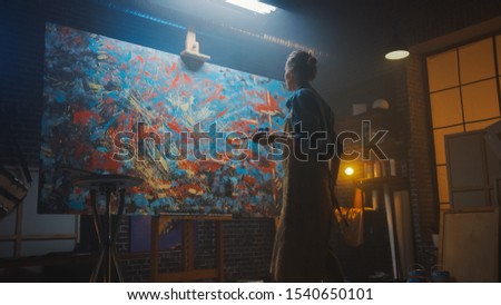 Female Artist Working on Abstract Oil Painting, with Broad Strokes of Paint Brush She Creates Modern Masterpiece. Dark Creative Studio Large Canvas Stands on Easel Illuminated, Tools Everywhere 