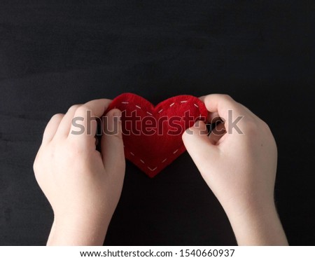 The child's hands hold a red cloth heart on a black background. Concept of love, baby love