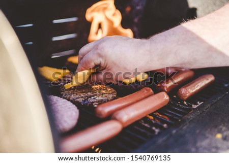 A closeup shot of a person putting cheese on the beef burger on the grill near hot dogs