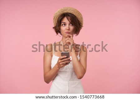 Thoughtful pretty young woman with casual hairstyle holding her chin with raised hand and looking aside pensively, keeping mobile phone in hands while standing over pink background