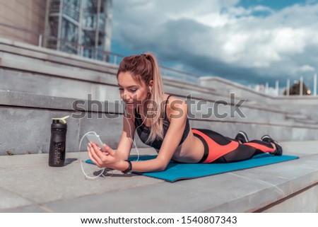 girl athlete lies on mat yoga gymnastics, looks phone, reads writes message, listens music headphones, rest workout summer day city Internet. Enjoys listens podcast lecture. Active lifestyle fitness