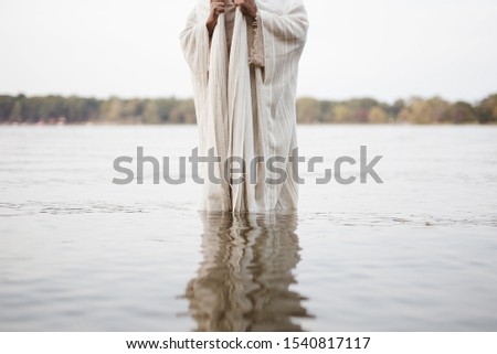 A person wearing a biblical robe standing in the water with a blurred background