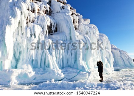 Baikal Lake. Small Sea Strait. A tourist photographs amazing ice crust with long icicles on the rocky shore of Olkhon Island on a sunny February day. Winter travel on the ice of a frozen lake
