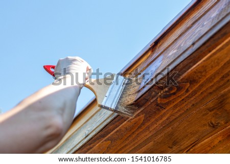 the worker painter smears with a brush with oil impregnation the wooden cladding of the house against the blue sky. Royalty-Free Stock Photo #1541016785