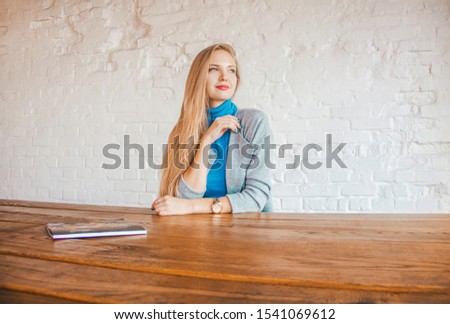 young casual girl with long blonde hair is sitting at a large wooden table on the white brick wall background, looking away and waiting somebody. lifestyle concept, free space, place for text