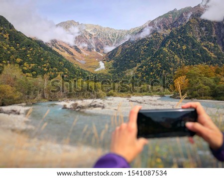Image of a blurry woman hand holding a black mobile phone to take a photo of a mountain with a stream on a sunny day