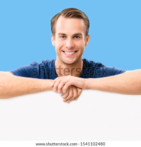 Happy smiling young man showing blank signboard with copy space area for slogan or some text, over blue color background. Square composition picture.