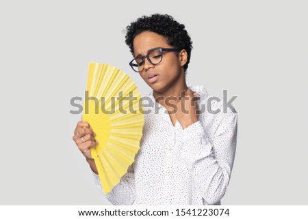 Head shot close up African American young woman in glasses waving fan, suffering from heat, sweaty exhausted girl cooling in hot summer weather, high temperature, isolated on grey background