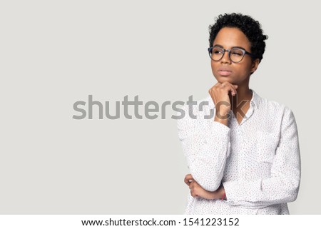 Thoughtful puzzled African American woman in glasses looking aside at at empty copy space, touching chin, customer pondering shopping offer, making decision, standing isolated on grey background Royalty-Free Stock Photo #1541223152