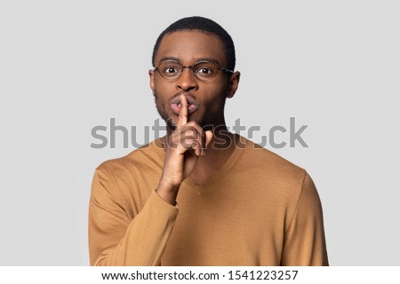 Head shot portrait close up funny African American man in glasses showing hush gesture isolated on grey studio background, keeping finger on lips, privacy, secret private information, sign of silence
