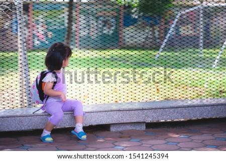 Cute bright Asian girl wearing a purple dress sitting on marble chair looking through the iron railings through to green lawn. There is a football gate in the lawn. Child aged 2 years and 8 months.