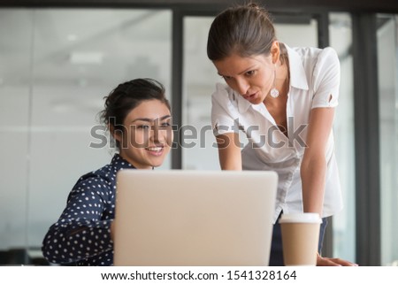 Young happy diverse female teammates working together on project at office. Senior manager mentor explaining software program to new employee or intern. Smiling indian worker showing report to boss. Royalty-Free Stock Photo #1541328164