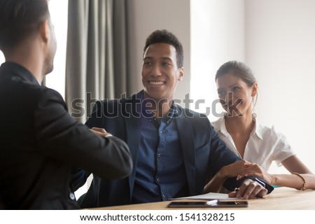 Smiling diverse couple shakes hands with real estate agent, lawyer, banker or life insurance manager after good deal. African american and caucasian family celebrating signing contract with advisor.