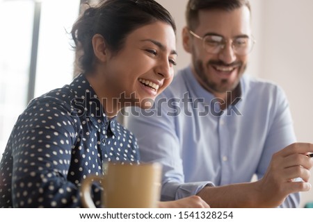 Happy young smiling diverse couple of female indian and male caucasian coworkers watching funny videos during coffee break, getting positive news, making good deal, holding video call with client Royalty-Free Stock Photo #1541328254