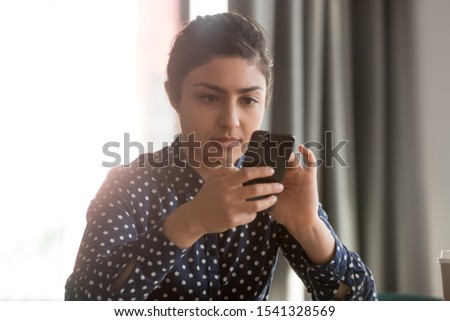 Concentrated young indian woman using cell phone virtual assistant, texting message in social media on cellphone, flipping news, checking email, viewing photos, using apps, reading articles at office