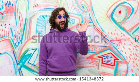young bearded crazy man feeling successful and satisfied, smiling with mouth wide open, making okay sign with hand against graffiti wall