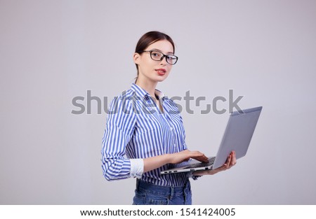 Close up, copy space. A girl manager looks straight in a striped white-blue shirt with black glasses and a laptop on left hand on a gray background. Employee of the year, business lady.