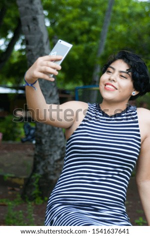 Relaxed young woman in makeup taking a picture of herself with her smart phone in the park, with green plants in the background. Concept of feminine beauty. Colorful photo
