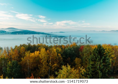 Aerial view to autumn foliage trees with misty fog, hill and blue sky in sunrise, Czech landscape, colored photo