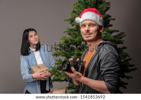 A young guy in a Christmas cap is filming on a mirrorless camera as a young girl with black hair is wrapping Christmas presents. Shooting for blog and vlog. Social Media Videos and Photos. DIY concept