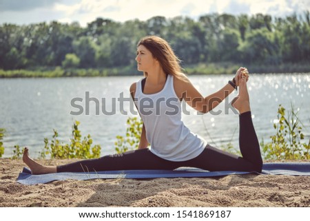 Woman in white undershirt and black leggings is practicing yoga performing yoga-asanas on a mat outdoors, near a riverside.