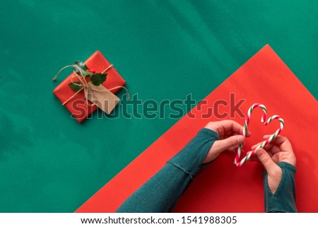 Creative diagonal geometric flat lay on green and red paper with long shadows of leaves. Hands in green t-shirt holding two stripy candy canes in shape of heart. Eco-friendly zero waste Christmas.