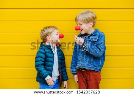 Red Nose Day Concept. Happy little brothers looks at each other wearing red clown noses having fun together on sunny autmn day outdoor, yellow background. Two kid playing together. Red Nose Day Royalty-Free Stock Photo #1542037628