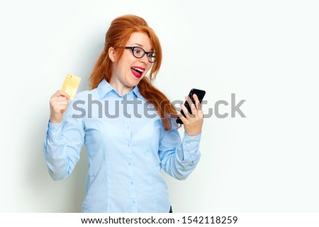 Portrait of an excited redhead girl looking at mobile phone while standing and holding credit card isolated over gray background copy space