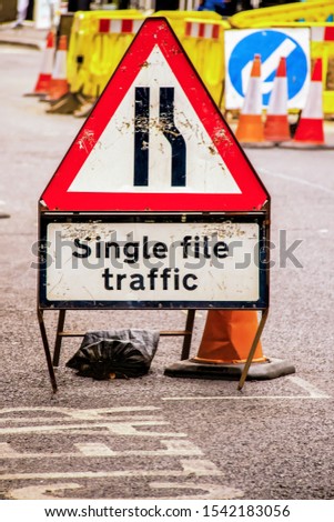 Single File Traffic - old grungy beat up traffic sign held in place by sand bag on pavement in front of barriers and traffic cones - UK - Selective focus